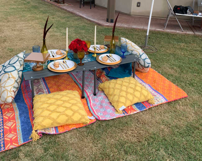 A colorful outdoor dining setup features a low table with plates, glasses, and flowers, surrounded by cushions on a patterned blanket under a decorative umbrella.