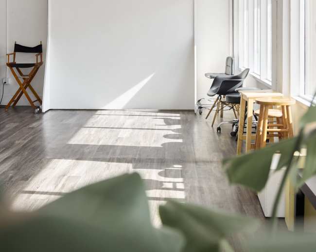 A bright, minimalist studio space features a large white backdrop, wooden flooring, and a few pieces of simple furniture, including a director's chair and a small stool, with natural light streaming through the windows.