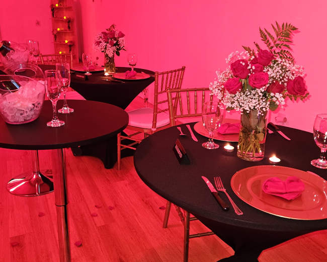The scene features elegantly set tables with black tablecloths, floral arrangements, and pink lighting in a decorated event space.