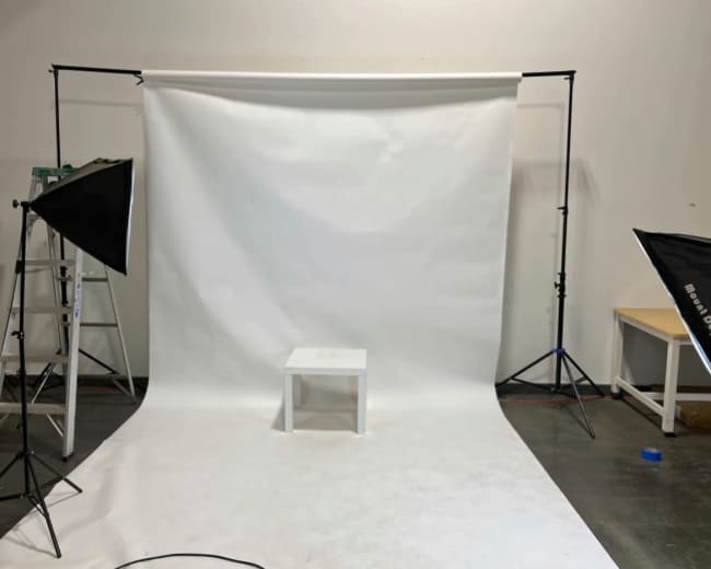 The image shows a photography studio setup with a white backdrop, a small white table in the center, and lighting equipment on either side.