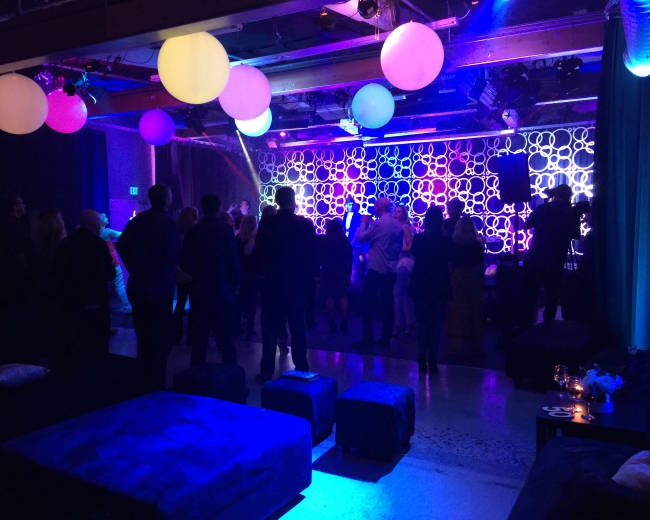A crowd of people dances under colorful hanging lights in a dimly lit event space.