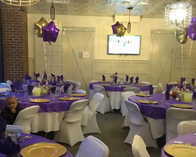 A banquet room with round tables set with purple tablecloths, gold plates, and decorative balloons, along with a television screen on the wall.