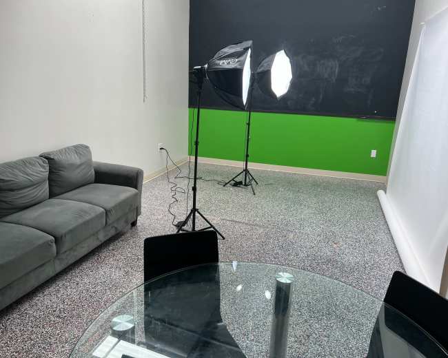 A minimal studio setup featuring a gray couch, a glass table, and a lighting umbrella in a room with a black chalkboard and a green accent wall.