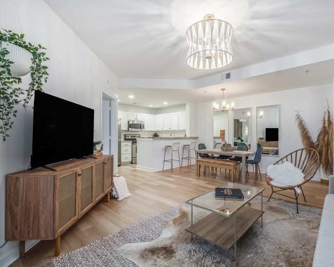 A modern living room features a television stand, a glass coffee table, a sectional sofa, and a dining area with a kitchen in the background.