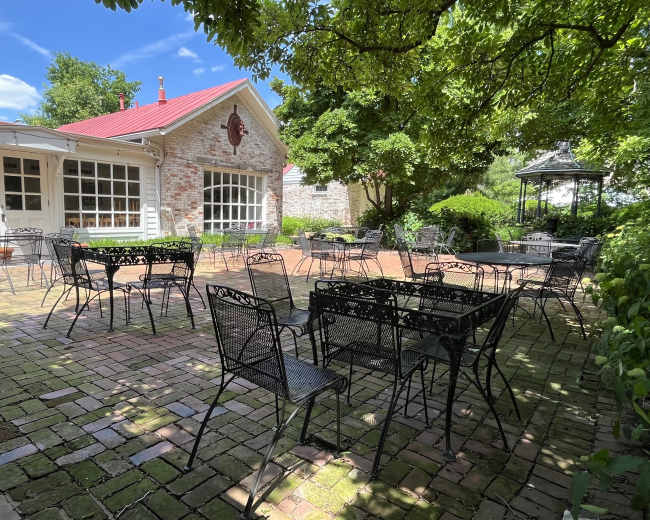 The image shows an outdoor seating area with several black wrought-iron tables and chairs arranged on a brick patio, surrounded by greenery and a building with large windows.