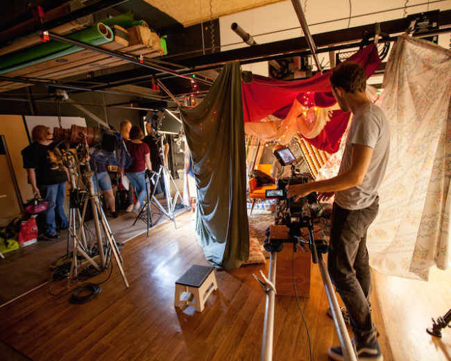 A film crew sets up equipment in a studio covered with colorful fabric drapes.