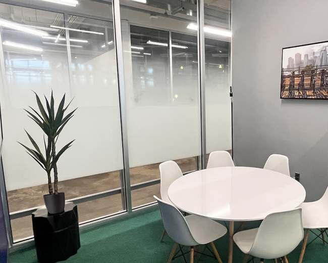 A round white table surrounded by six chairs is set in a glass-walled conference room featuring a plant and a cityscape painting on the wall.