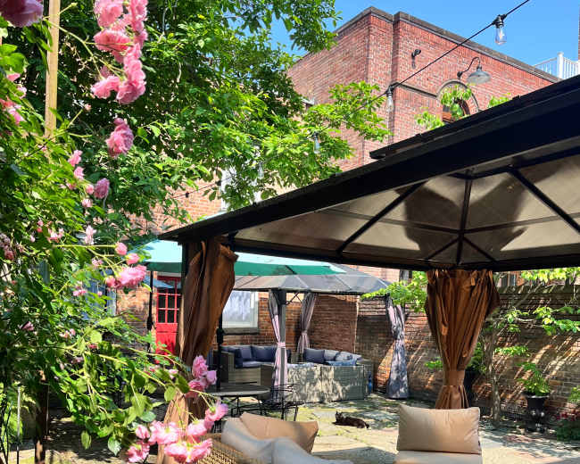 The image shows a cozy outdoor seating area surrounded by greenery and blooming flowers, with a covered pavilion and patio furniture.