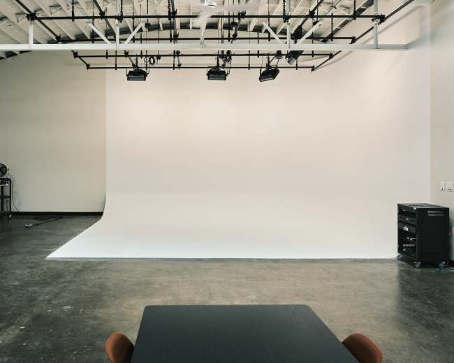 The image shows a spacious studio with a white backdrop, overhead lighting, and a black table surrounded by chairs in the foreground.