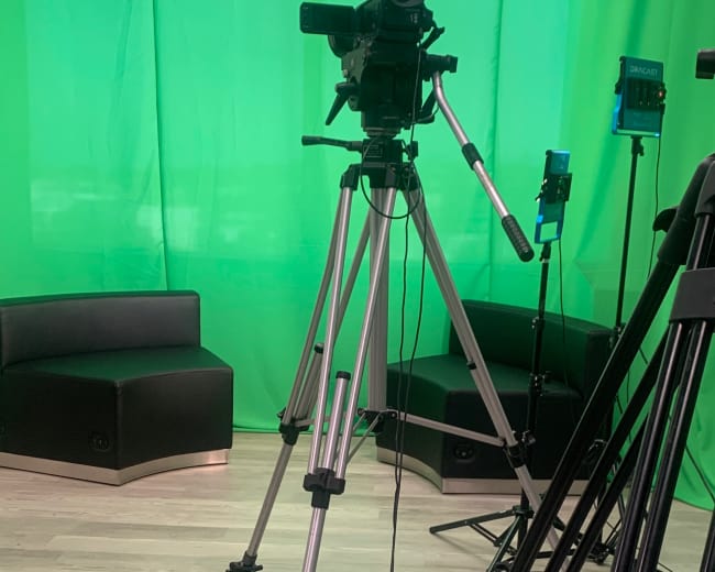 The image shows camera equipment set up in front of a green backdrop in a studio environment.