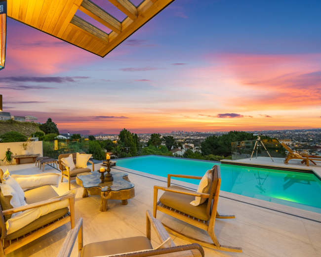 A modern outdoor patio features comfortable seating around a table, overlooking a swimming pool with a sunset view of the city skyline.