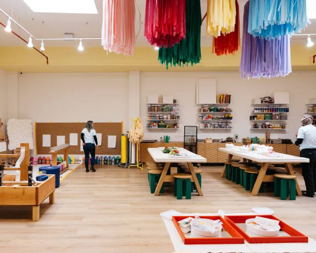A brightly lit art studio features large wooden tables, colorful decor hanging from the ceiling, and various art supplies organized on shelves.