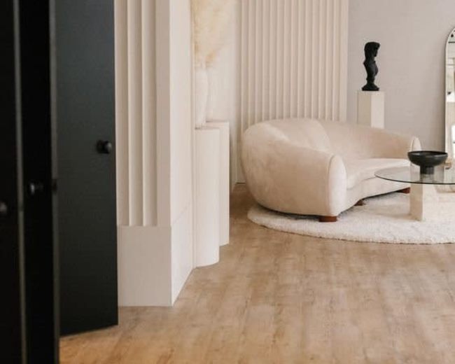A modern interior featuring a curved beige sofa, a round glass coffee table, and minimalist decor against textured white walls.