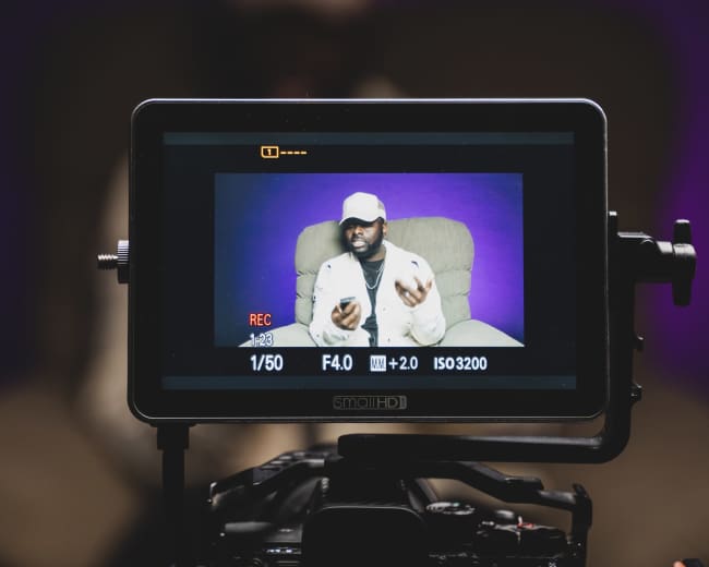 A camera displays a person gesturing while being recorded against a purple backdrop.