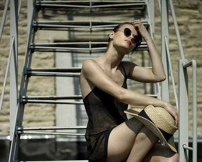 A woman in a black dress sits on metal stairs while holding a straw hat and wearing sunglasses.
