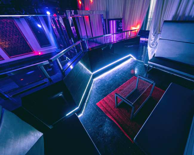 The image shows a modern lounge area featuring black seating, a red rug, and ambient lighting in a dimly lit environment.