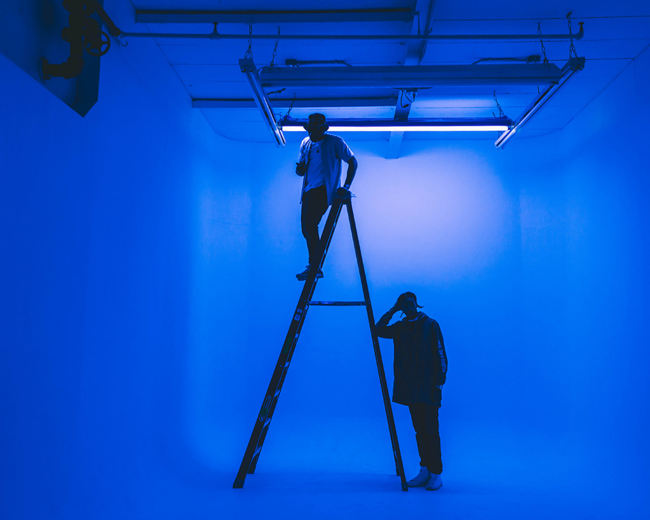Two individuals are positioned in a blue-lit studio, one standing on a ladder and the other on the ground beside it.