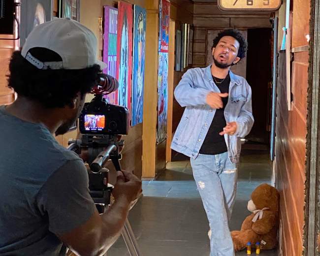 A person wearing a denim jacket performs in a hallway while another individual records the scene with a camera set on a tripod.