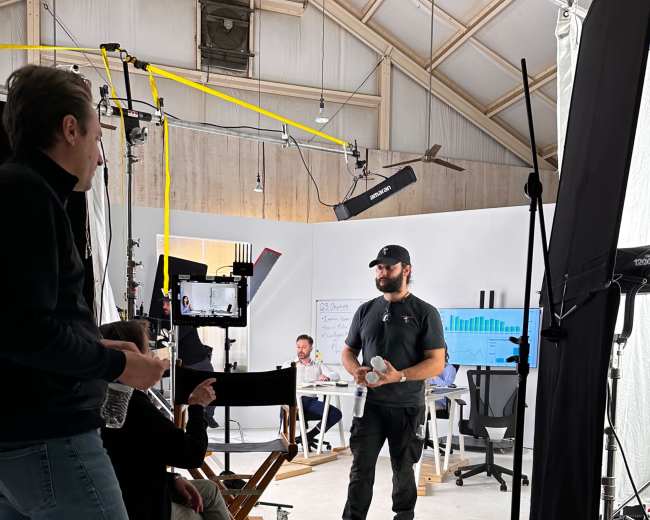 A production team is set up in a studio, with one person standing and presenting to a small audience seated nearby, while various cameras and lighting equipment are arranged around the space.