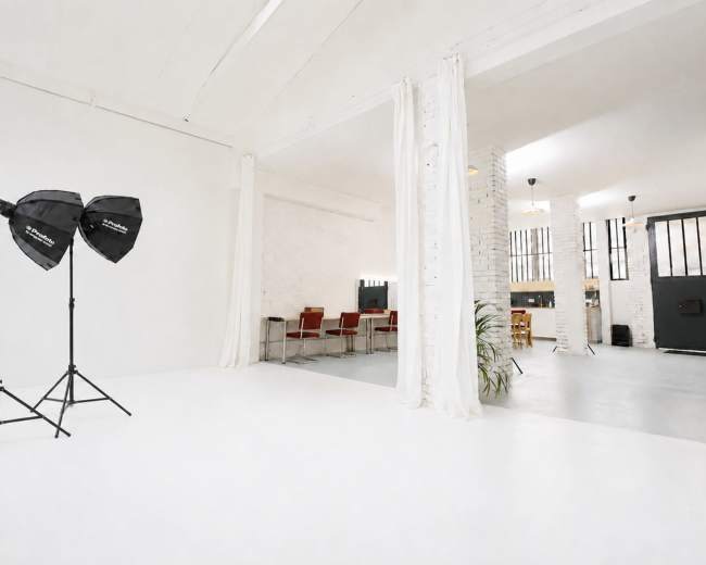 A bright studio space features two light stands with softboxes, white walls, and an area with red chairs and a dining table in the background.