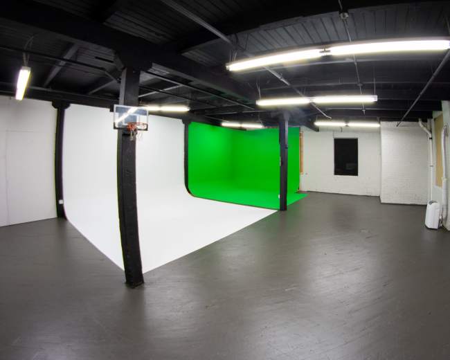 A spacious studio with a white wall and a green screen backdrop, featuring black beams and fluorescent lighting.