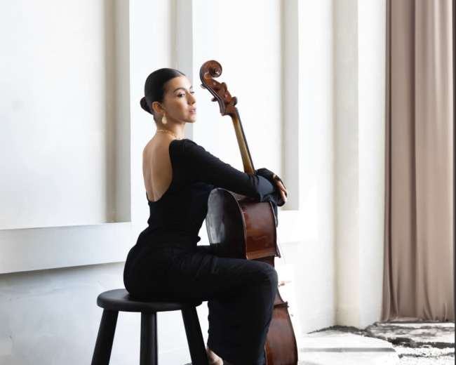 A woman dressed in black sits on a stool, elegantly posed with a cello beside her in a minimalist interior setting.