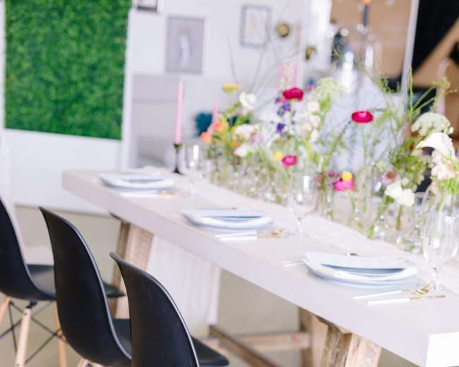 A long dining table is set with plates and glassware, surrounded by black chairs with wooden legs, and decorated with a floral arrangement.