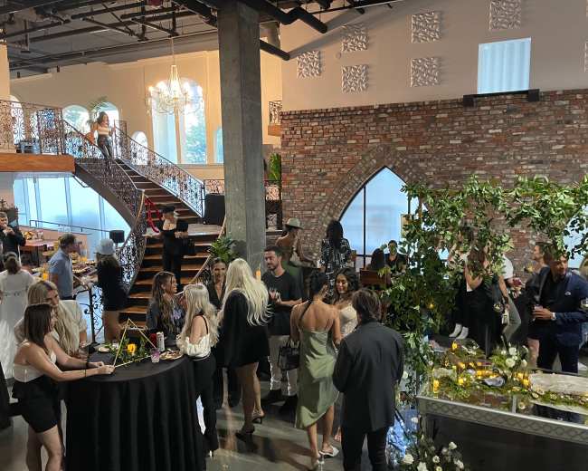 A group of people socializes at a stylish event held in a modern venue with exposed brick walls and elegant architecture.