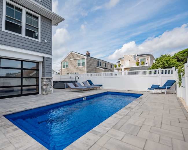 A rectangular swimming pool surrounded by lounge chairs and a fenced patio area beside a house.