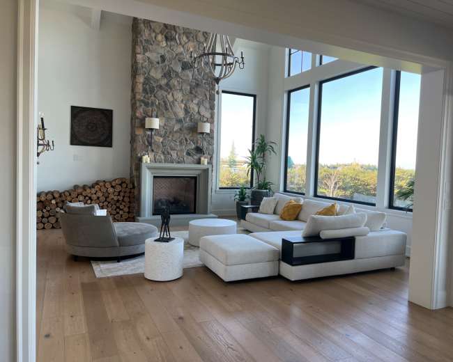 The image shows a modern living room featuring large windows, a stone fireplace, a sectional sofa, and a circular coffee table.