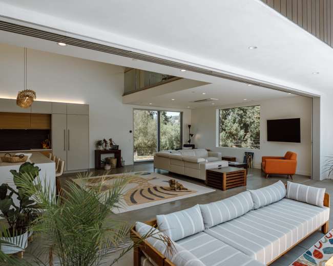 The image shows a modern living room with a spacious layout, featuring a sectional sofa, an armchair, a coffee table, and large windows that provide natural light.