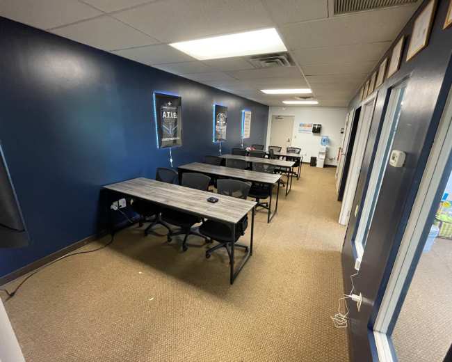 The image shows a long, narrow room with several gray tables and black chairs arranged in rows along a dark blue wall.