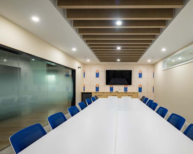 A modern conference room features a long white table surrounded by blue chairs, with a decorative wall and a television at one end.