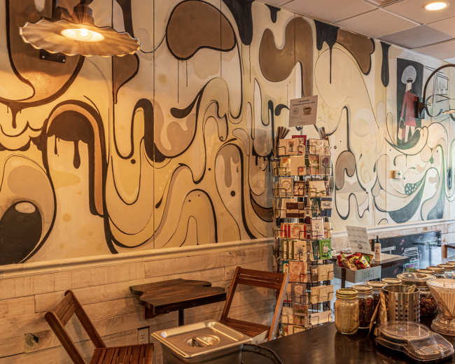 The image shows a coffee shop interior with a colorful abstract mural on the wall, wooden tables, and a display of packaged goods.