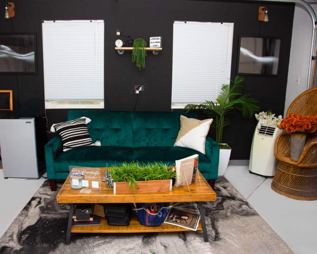 The image shows a living space with a green velvet couch, a wooden coffee table on a patterned rug, and various decorative items against a dark wall.
