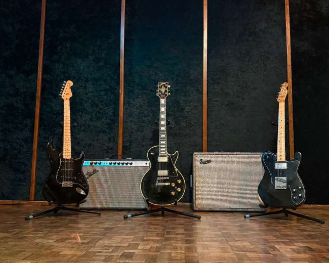 The image shows four guitars arranged in front of amplifiers on a wooden floor against a dark background.