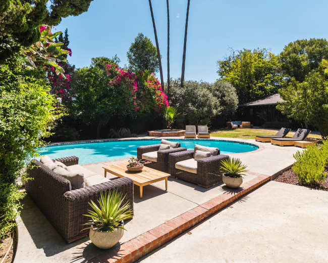 The image shows a backyard featuring a swimming pool surrounded by patio furniture, palm trees, and colorful vegetation.