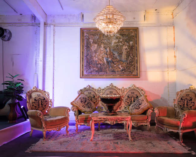 The scene features a vintage seating arrangement with ornate chairs and a matching table, set against a wall adorned with a large framed painting and a chandelier overhead.