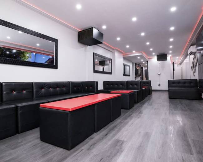 The image shows a modern interior of a lounge with black leather seating, a red table in the center, and ambient lighting.