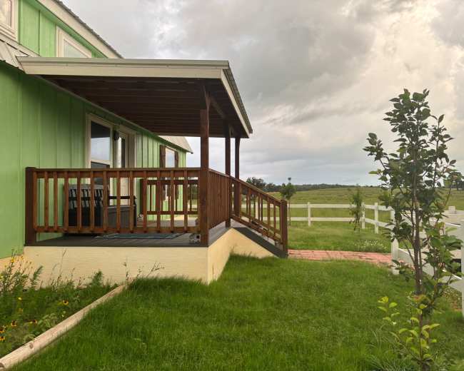 A green building with a wooden deck features a sloped entrance and is surrounded by grassy landscaping and a white fence.