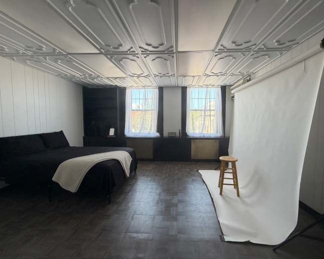 The image shows a spacious room with a black bed, a white backdrop on a stand, and large windows letting in natural light.
