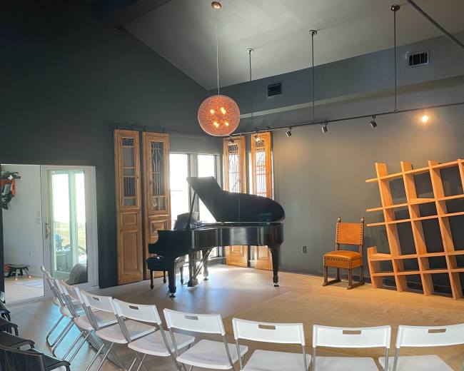 A grand piano is positioned on a wooden stage, surrounded by rows of black and white chairs in a room with dark walls and large windows.