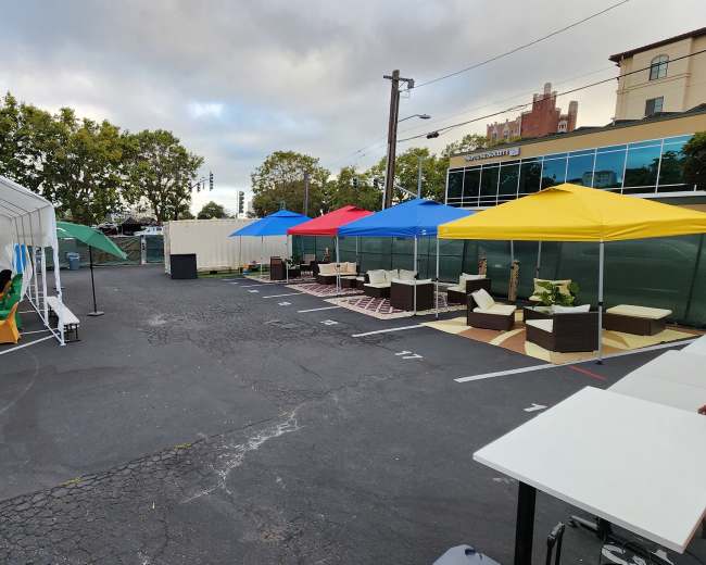 The image shows a parking lot divided by colorful canopies, with seating arrangements set up on either side.