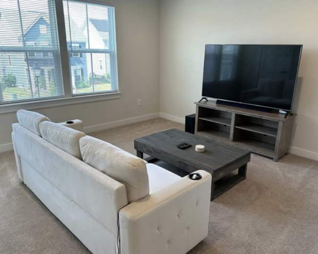 A sunlight-filled living room features a white couch, a wooden coffee table, and a large television on an entertainment unit.