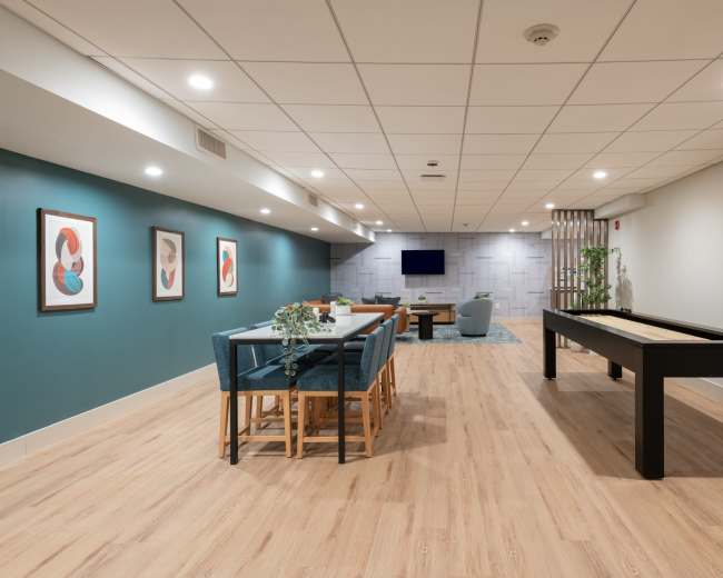 The image shows a spacious, modern game room featuring a pool table, a dining area with seating, and a large flat-screen TV on the wall.