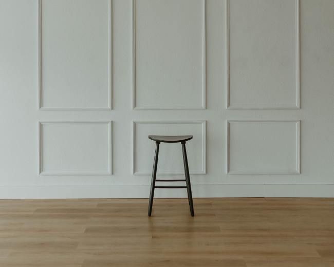 A black stool stands against a plain white wall with decorative paneling.