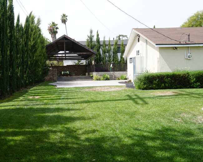 A neatly trimmed lawn bordered by tall, slender trees, with a covered patio area and a house visible in the background.