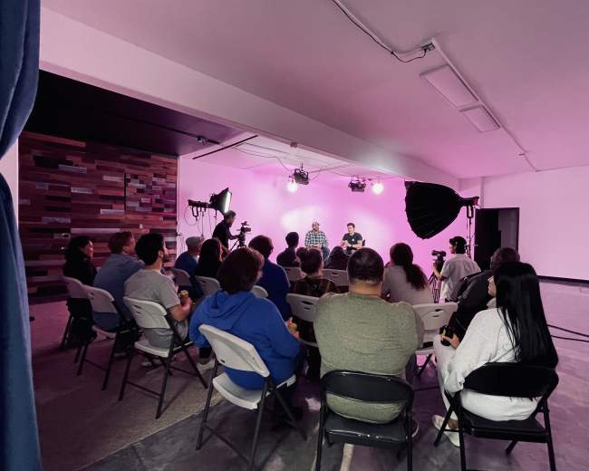 A group of people sits in folding chairs facing two individuals being filmed in a brightly lit studio space.