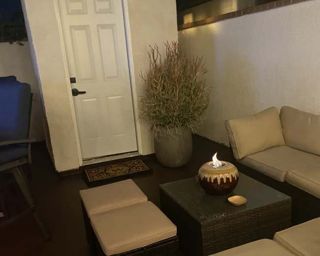 A small outdoor seating area features a couch, two ottomans, and a decorative pot with a plant, adjacent to a closed white door.