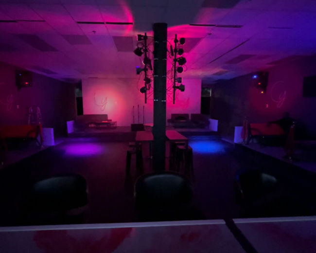 The image shows an empty nightclub interior with dim lighting and a stage featuring a spotlight setup.